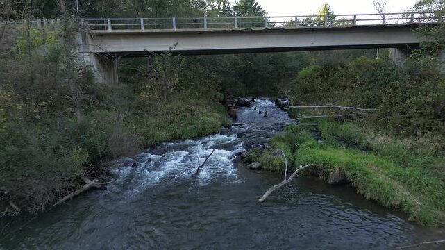 Passing under a bridge spanning serene flowing rapids. Water tumbles beneath an overpass in a rural countryside. Green vegetation and forest surround the aged infrastructure. Aerial Drone footage