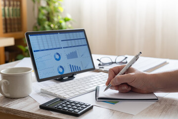 Fototapeta premium Person working from home, reviewing financial reports and accounting data on a tablet, calculating figures with a calculator, and writing notes in a notebook, managing budgeting tasks