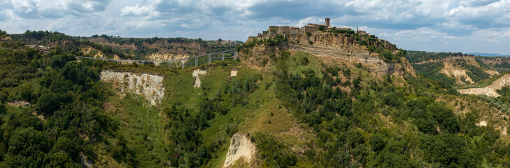 Obraz premium Aerial front view of Civita di Bagnoregio, Italy, showing medieval stone buildings and a tall bell tower atop a rocky cliff, with the entrance gate and bridge path visible against distant green hills.