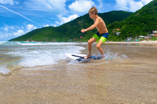 Young boy running, jumping on skimboard and sliding by shallow water on sunny sea beach. Skimboarding, childhood adventure, active lifestyle, watersports, summer vacation and fun outdoor activities.