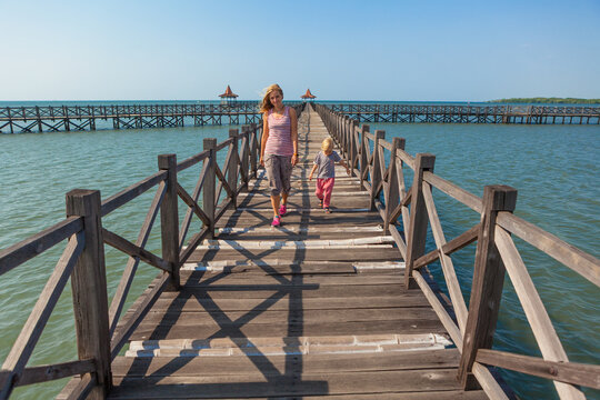 Family with children walks along a long wooden pier stretching into the blue sea on sunny day. Clear blue sky, calm ocean create peaceful, tropical atmosphere. Travel with kids, summer, beach vacation