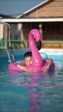 Young boy relaxes on pink flamingo inflatable in outdoor swimming pool on sunny summer day, eyes closed, peaceful leisure moment