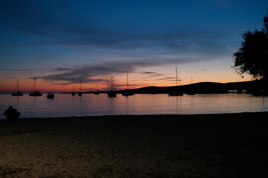 Sunset blue hour over Paralia Parikia, Paros, Greece, with sailboat silhouettes reflected on calm Mediterranean waters beneath a colorful twilight sky.
