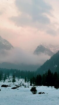 4K Vertical view of snowfall in beautiful mountain valley during sunset at Kashmir, India. Orange sunset clouds in sky and snow covered mountains at Aru Valley during winter season. Dramatic nature la