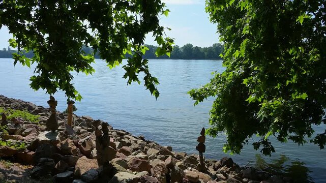 Steine auf einem Flussufer bei strahlendem Sonnenschein und ruhigem Wasser, umgeben von gr&uuml;nem Laub, Rhein, Eltville am Rhein, Rheingau, Hessen, Deutschland
