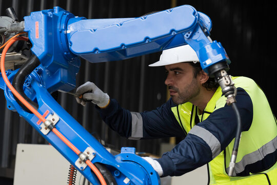 Male engineer wearing uniform, white hard hat and gloves repair automatic robotic arm for welding. Male technician inspecting quality automatic welding robot arm machine with hand tools