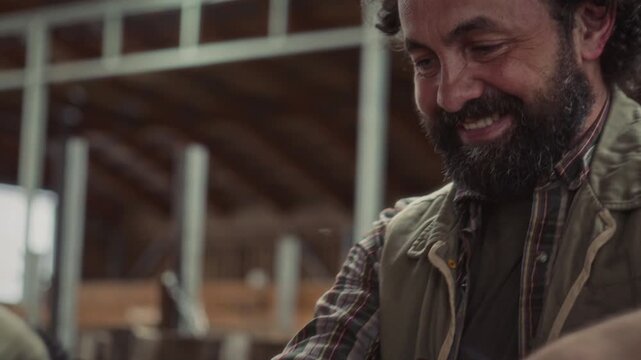 Happy bearded male farmer petting black horned cow with affection and smiling as taking care of livestock inside barn