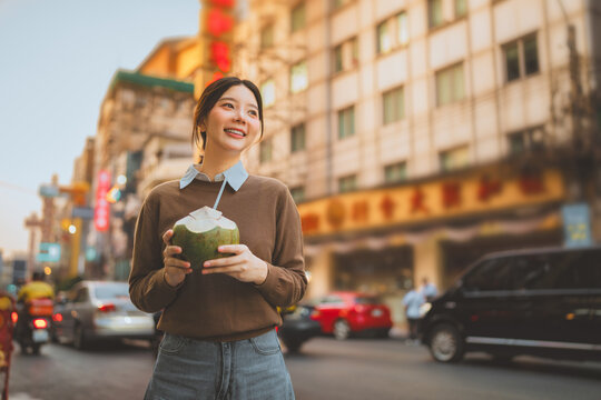 Happy young Asian woman traveler holding fresh coconut drink at chinatown street market Bangkok looking away with cheerful smile tourist exploring local food and culture urban lifestyle, Thailand