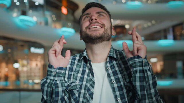 Happy Caucasian man crossing fingers waiting result smiling wishing anticipation winning in shopping mall positive business male client businessman wait wish guy hope smile dreaming success indoors