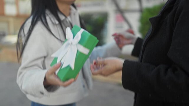 Woman smiles with eyes closed and hugs her friend who holds a green gift box tied with a white bow on a street outside a building; joy friendship warmth.