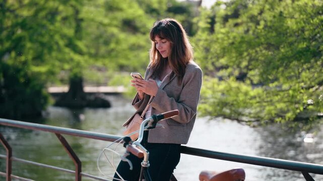 Video of attractive woman using her smartphone while leaning on the railing beside the lake in the park.