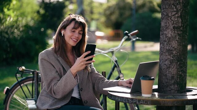 Video of beautiful woman working on a laptop while using smartphone in the park.
