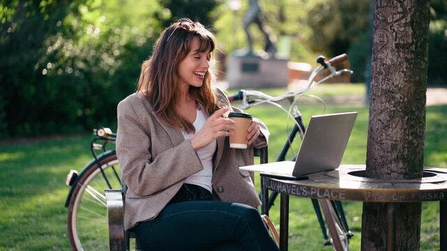 Video of beautiful woman doing videocall with laptop while drinking coffee in the park.
