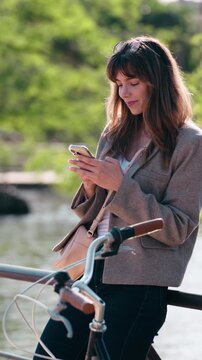 Video of attractive woman using her smartphone while leaning on the railing beside the lake in the park.