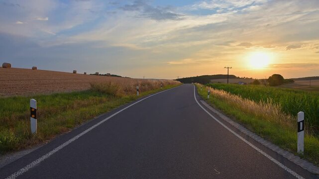 Eine Landstra&szlig;e, die sich durch die Felder schl&auml;ngelt, w&auml;hrend die Sonne am Horizont untergeht, Sommer, Reichartshausen, Amorbach, Odenwald, Bayern, Deutschland