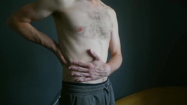 Shirtless caucasian man with a hairy chest rubbing his belly with his hands because he is feeling abdominal pain, possibly due to indigestion, gastritis, or colic in a dark room