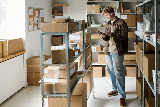 Young adult man checking inventory on tablet in stockroom, managing parcel fulfillment and warehouse workflow. Ideal for e commerce logistics, small business operations, order processing
