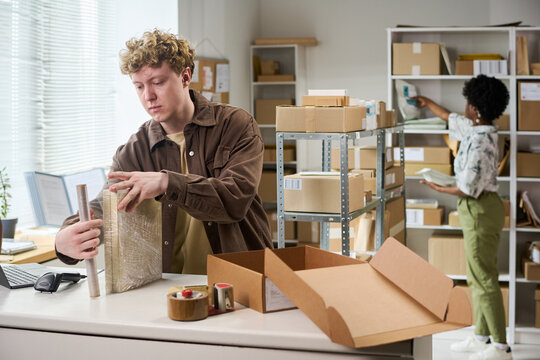 Young Caucasian man packing parcel with stretch wrap while Black woman organizing inventory on shelves in small warehouse. Supporting ecommerce fulfillment, order processing, retail logistics