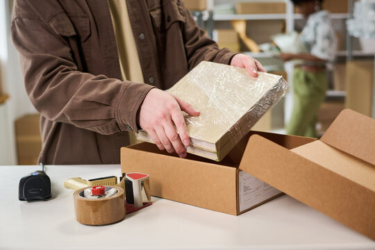 Close up of Caucasian young adult packing wrapped books into shipping box, preparing retail order at fulfillment workspace. Ideal for ecommerce, order processing, small business