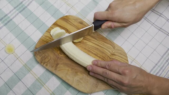 Woman's hands using knife to cut banana into slices on cutting board - high shot