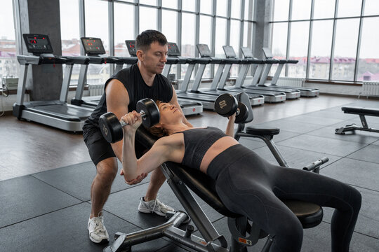 Personal trainer spotting woman performing dumbbell chest press on gym bench in bright fitness studio with cardio machines and focused strength training atmosphere