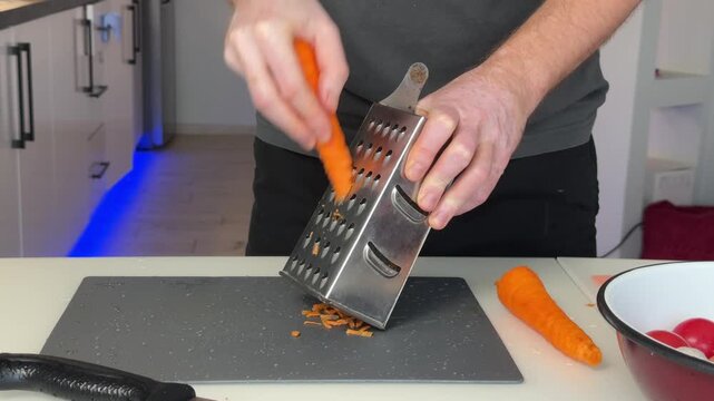Hands grating peeled carrot over dark cutting board with stainless box grater in modern kitchen, producing bright orange shreds and scattered fragments during meal preparation