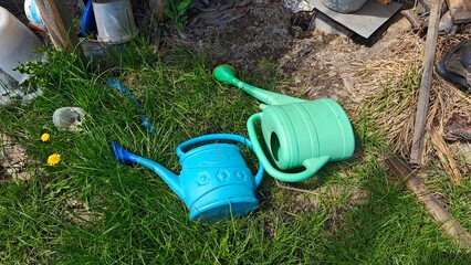 Two plastic watering cans, one blue and one green, lying on grass in a garden, surrounded by dandelions and garden debris in a rural outdoor setting © Olena