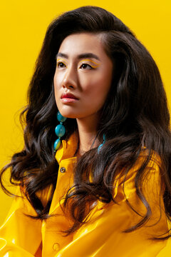 Asian woman in yellow raincoat wearing blue stone earrings and creative eyeliner looking away against bright background