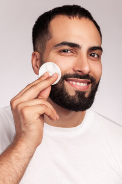Smiling man applying skincare lotion with cotton pad wiping bearded face for hygiene wearing casual white t-shirt posing isolated over gray background