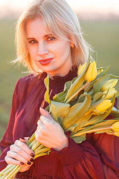 Blonde woman holding yellow tulips near her face while looking at camera in field during sunset with soft lens flare