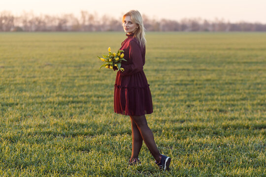 Woman with short blonde hair in tiered burgundy ruffled dress posing in green field during sunset while holding a bouquet of yellow tulips in hands and looking at camera