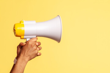 Profile portrait of astonished worker wearing blue overalls and red hat holding megaphone in hands and looking at camera with widely open mouth. Indoor studio shot isolated on yellow background. © khosrork