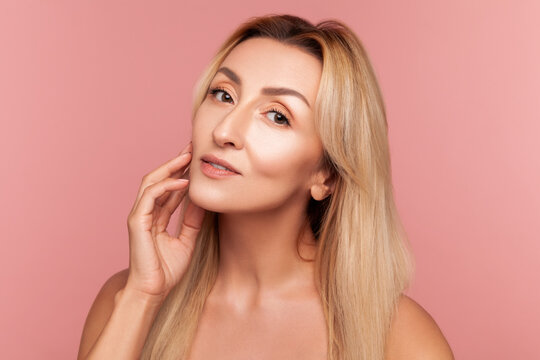 Beautiful adult woman touching her smooth face skin gently for aesthetic dermatology and rejuvenation campaign isolated on pink background