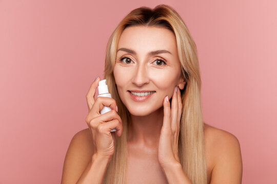 Smiling woman holding white serum bottle for face hydration and anti-aging treatment during daily cosmetic beauty routine isolated on pink background