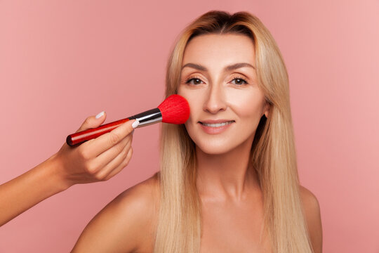 Blonde female model getting makeup with large red brush professional artist enhancing skin tone and beauty isolated on pink background