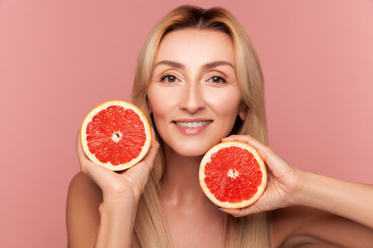 Happy woman holding two grapefruit halves for skin health and vitamin C infusion beauty procedure isolated on pink background