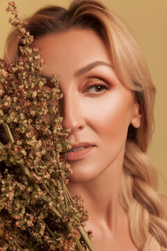 Beautiful female face partially hidden by dried plants while looking away showing bronze eye makeup isolated over yellow background