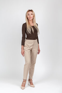 Full length shot of blonde woman standing in high heels wearing brown long sleeve shirt and beige trousers looking at camera isolated over gray background