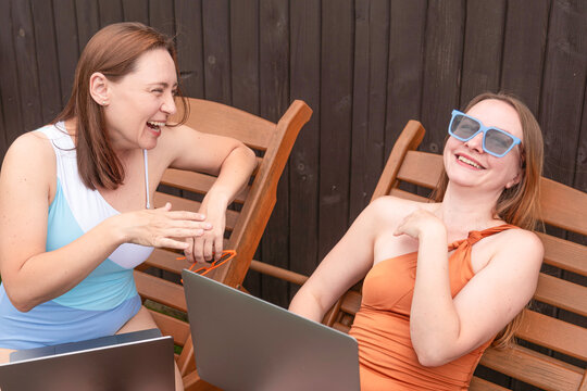Two women laughing on deck chairs with laptops outdoors