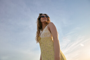 Obraz premium Low Angle View of Young Woman in Yellow Summer Dress and Sunglasses Outdoors Against Blue Sky