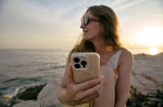 Young Woman Holding Smartphone at Rocky Sea Coast During Sunset