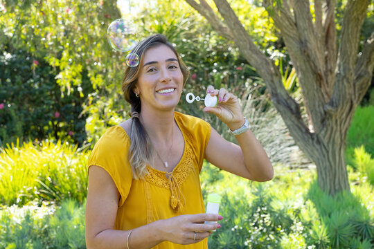 Adult female wearing mustard blouse holding white bubble wand and bottle, blowing bubbles in garden