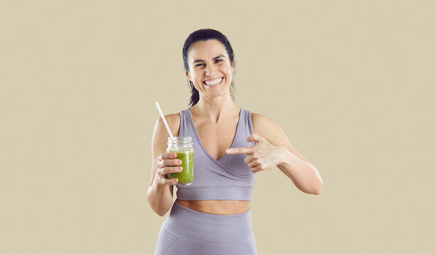 Smiling woman athlete pointing at healthy smoothie. Studio portrait in sportswear with smoothie, supporting clean eating on neutral background. Theme healthy fitness lifestyle and nutrition.