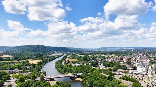 Vogelperspektive auf Saarlouis mit der vorbeiflie&szlig;enden Saar, einer markanten Stra&szlig;enbr&uuml;cke, dichter gr&uuml;ner Vegetation und der Stadtlandschaft unter blauem Wolkenhimmel.