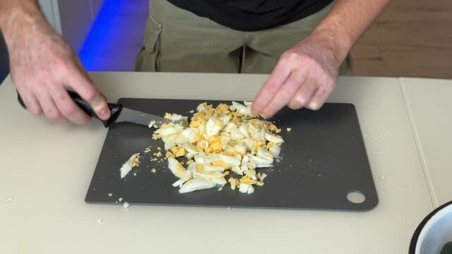 Hands chopping hard boiled eggs on dark cutting board in bright home kitchen overhead view showing meal prep, food preparation, cooking process, and scattered egg pieces