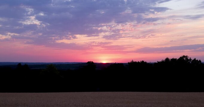 Real-time cinematic footage of the sun setting beyond a rural field beneath textured evening clouds. Atmospheric establishing shot conveying the transition from day to night.