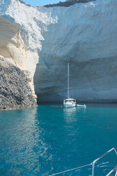 White sailboat resting in crystal-clear turquoise water beneath the towering white cliffs and sea caves of Kleftiko, Milos, Greece in a sunny summer day
