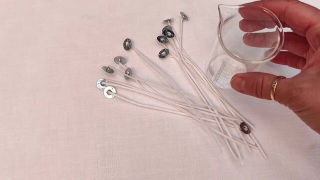 Woman places a glass measuring cup beside candle wicks on a white tablecloth, candle making supplies, handmade production, DIY craft, creative workspace, flat lay.
