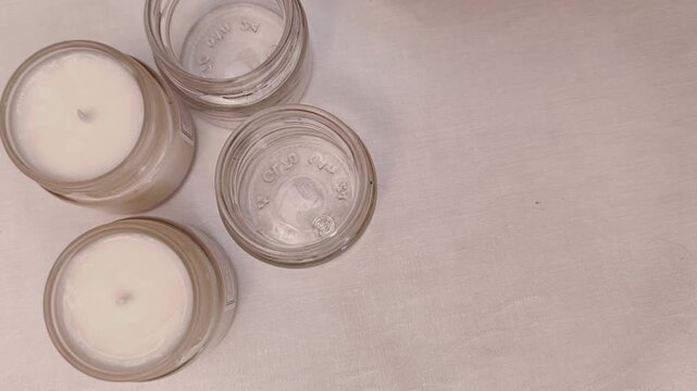 Woman places an empty glass candle jar beside finished soy candles on a white tablecloth, candle making process, handmade production, artisan workshop, minimalist style.
