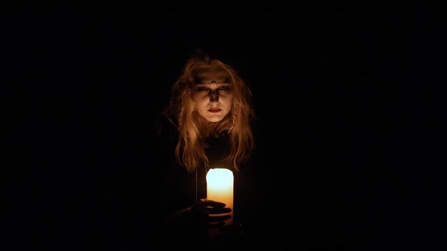 Centered close up shows person holding tall lit candle at chest level, facing camera in dark setting with warm glow, strong shadow contrast, and eerie ceremonial atmosphere, with candle, portrait